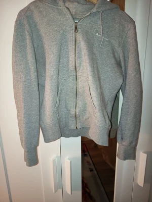 Grå zip-hoodie från Champion - Grå hoodie med dragkedja från Champion. Klassisk modell med huva, kängurufickor och ribbade muddar. Liten Champion-logga på bröstet och ärmen. Perfekt för en avslappnad och sportig stil.