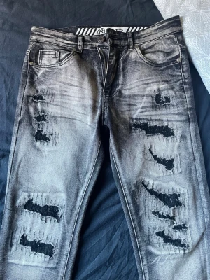 Grå slitna jeans från RGSTR - Säljer ett par grå jeans från RGSTR med tydliga slitningar och destroyed detaljer på både fram- och baksida. Jeansen har fem fickor, knappgylf och cool tvättad look. Materialet är 80% bomull, 15% polyester och 5% elastan för skön stretch.