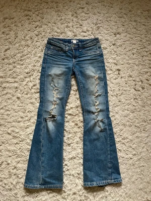 Blå bootcut jeans med slitningar - Snygga blå bootcut jeans med kraftiga slitningar och hål längs benen. Jeansen har en klassisk femficksmodell och är tillverkade i denim med en tvättad finish. Perfekta för en avslappnad och trendig look.