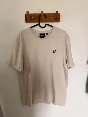 Beige t-shirt från Lyle & Scott - Snygg beige t-shirt från Lyle & Scott med klassisk rund hals och ikonisk gul broderad logga på bröstet. Tillverkad i mjuk bomull som känns skön mot huden. Perfekt för en clean och stilren look.