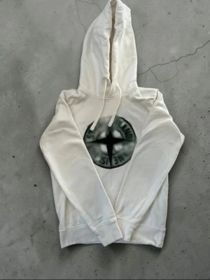 Vit hoodie från Stone Island - Snygg vit hoodie från Stone Island med klassisk logga i svart och grönt på bröstet. Hoodien har dragsko i huvan och ribbade muddar vid ärmslut och nederkant. Perfekt för en avslappnad och trendig stil.