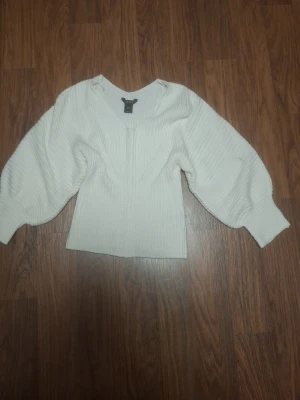 Vit blus med ballongärmar från Lindex - Snygg vit blus från Lindex med trendiga ballongärmar och v-ringning. Blusen har en ribbad struktur och är figurnära i midjan vilket ger en modern siluett. Perfekt att styla med jeans eller kjol för en cool look.