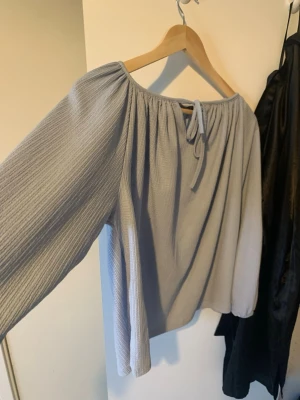 Ljusgrå offshoulder-blus från 157 - Superfin ljusgrå blus från 157 med offshoulder-modell och långa ärmar. Blusen har en rynkad resår upptill och knytband framtill. Materialet är lätt och har en diskret struktur som ger en avslappnad vibe.