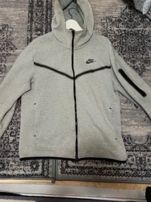 Grå Nike Tech Fleece hoodie (M) Helt NY - Snygg grå Nike Tech Fleece hoodie med hel dragkedja och svarta detaljer. Har huva, två fickor fram och en extra ficka med dragkedja på ärmen. Perfekt för en sportig och avslappnad stil. Nike-logga på bröstet och modern passform.