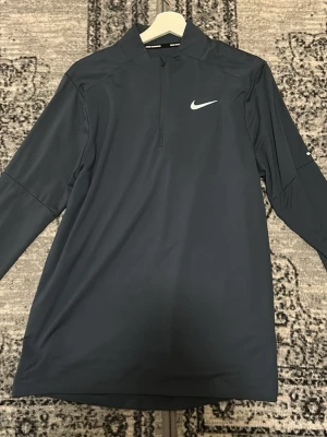 Mörkblå långärmad träningströja Nike - Sportig mörkblå långärmad träningströja från Nike med halv dragkedja framtill och vit logga på bröstet. Perfekt för träning eller uppvärmning. Tillverkad i ett syntetmaterial som andas.
