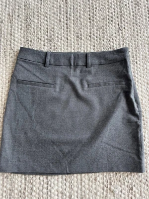 Mörkgrå minikjol från H&M  - Stilren mörkgrå minikjol med bälteshällor och två dekorativa fickor bak. Kjolen har en klassisk form och är perfekt för en clean och enkel look. Tillverkad i ett mjukt material som känns bekvämt hela dagen.