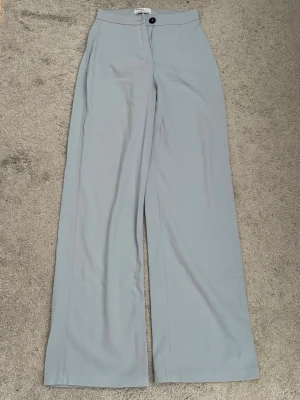 Ljusblå vida kostymbyxor Bershka - Snygga ljusblå kostymbyxor från Bershka med hög midja och vida ben. Byxorna har klassisk knapp och dragkedja framtill och är perfekta för en clean och stilren look. Tillverkade i ett mjukt och lätt material som känns bekvämt hela dagen.