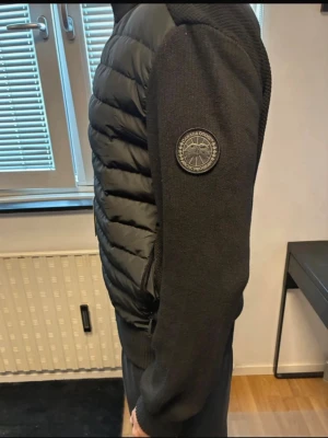 Svart cardiganjacka från Canada Goose - Snygg svart cardiganjacka från Canada Goose med quiltad front och stickade ärmar. Jackan har en diskret patch på ärmen och dragkedja framtill. Perfekt för lager-på-lager och ger en clean look till din outfit.