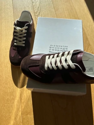 Maison Margiela vinröda sneakers - Snygga sneakers från Maison Margiela i vinrött läder och mocka med vita snören. Klassisk låg modell med stilren design och diskreta detaljer. Perfekta för dig som vill ha något unikt och trendigt i garderoben.