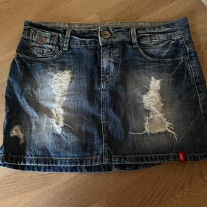 Blå jeanskjol med slitningar - Säljer en blå jeanskjol med coola slitningar framtill och klassiska fickor både fram och bak. Kjolen har knapp och dragkedja i midjan samt en liten röd detalj vid nederkanten. Perfekt för en avslappnad och trendig look.