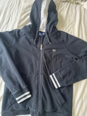 Marinblå kofta från Fred Perry - Marinblå kofta från Fred Perry med dragkedja, vit logga på bröstet och gråvita ränder på muddar och nederkant. Huva med vit meshfoder och dragsnören. Lite stor men skön på mig som oftast har S. Bra skick och skönt material.