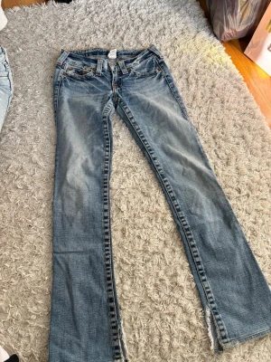 Blå bootcut jeans från true religion med slitningar - Säljer ett par blå bootcut jeans med slitningar och råa kanter nertill. Jeansen har klassisk femficksdesign och ljus tvätt med snygga detaljer. Perfekta för dig som gillar en avslappnad och trendig stil.