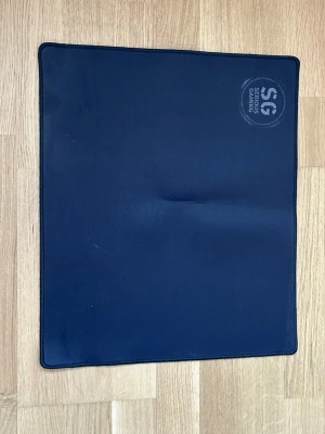 Svart musmatta Serious Gaming - En svart, rektangulär musmatta från Serious Gaming med rundade hörn och logga tryckt i ena hörnet. Tillverkad i mjukt tygmaterial med sydda kanter för extra hållbarhet. Perfekt för gaming eller kontorsbruk.