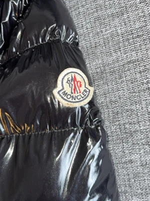 Svart glansig dunjacka från Moncler - Säljer en svart glansig dunjacka från Moncler med klassisk logotyp på ärmen. Jackan har quiltad design och är tillverkad i ett vattenavvisande material med dunfyllning som håller dig varm under kalla dagar. Perfekt för vintern och riktigt snygg streetstil.
