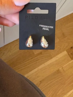 Pilgrim örhängen med sötvattenspärlor - Säljer ett par droppformade örhängen från Pilgrim med skimrande sötvattenspärlor i silverton. Örhängena har en modern och stilren design som ger en elegant touch till din look. Perfekta för dig som gillar unika accessoarer med naturliga material.