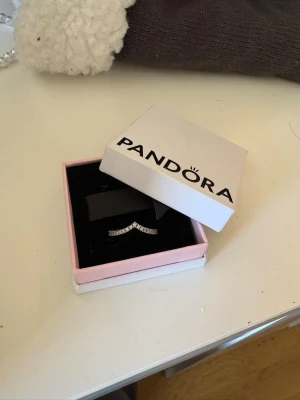 Silverring med stenar från Pandora - Säljer en snygg silverfärgad ring från Pandora med gnistrande stenar i en elegant vågig design. Ringen kommer i original ask och har en modern look som passar perfekt till din smyckessamling.