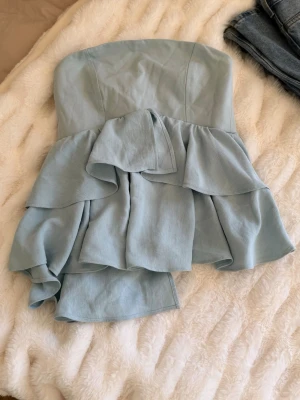 Ljusblå axelbandslös volangtop - Supersöt ljusblå axelbandslös topp med volanger och bandeau-modell. Toppen har en flowy passform och är perfekt för dig som vill ha en feminin och trendig look. Tillverkad i ett mjukt tyg som känns skönt mot huden.