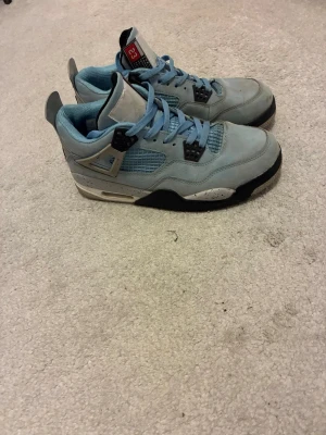 Jordan 4 University Blue sneakers - Snygga Jordan 4 University Blue sneakers i ljusblå mocka med svarta och grå detaljer. Skorna har meshpaneler på sidorna, svart Jumpman-logga på hälen och vita sulor med stänk. Klassisk siluett med högt skaft och coola detaljer.