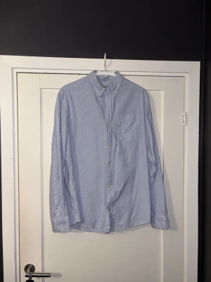 Blårandig skjorta från Jack & Jones - Klassisk blårandig skjorta från Jack & Jones med vit bas och tunna blå ränder. Skjortan har lång ärm, knappar framtill och en bröstficka. Perfekt för en clean och stilren look.