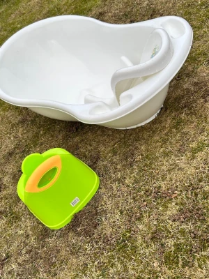 Vit babybadbalja och grön potta - Säljer en vit babybadbalja i plast med inbyggt stöd och ett gulligt djurmärke, samt en grön och orange potta i plast. Perfekt för småbarn och enkel att rengöra. Båda produkterna är lätta att bära och använda hemma.