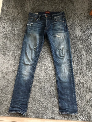 Blå slitna jeans från Jack & Jones - Säljer ett par blå jeans från Jack & Jones med slitningar och trasiga detaljer på benen. Jeansen har en smal passform, klassisk femficksmodell och är gjorda i denim med snygg tvättad look. Perfekta för en avslappnad och trendig stil.