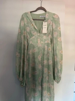 Grön blommig långklänning Adoore - Långklänning från Adoore i ljusgrönt med blommigt mönster i vitt och gult. Klänningen har v-ringning, långa ärmar och knappar framtill. Tillverkad i ett lätt och luftigt material, perfekt för vår och sommar. Helt ny, aldrig använd med prislapp kvar!🌸