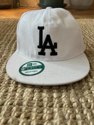 Vit LA-keps från New Era - Snygg vit keps från New Era med svart LA-logga framtill. Modellen är 9FORTY och har justerbar snapback baktill för perfekt passform. Klassisk böjd skärm och broderade detaljer. Perfekt accessoar för en sportig och trendig look.