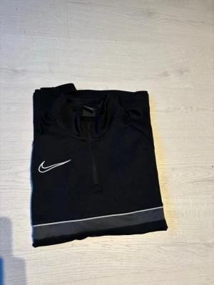 Svart Nike träningströja med dragkedja - Snygg svart långärmad träningströja från Nike med vit logga på bröstet och en diskret grå/vit rand över bröstet. Tröjan har halv dragkedja framtill och är tillverkad i ett lätt och funktionellt material som passar perfekt till träning.