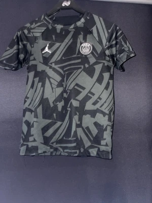 PSG x Jordan svart mönstrad t-shirt - Snygg svart t-shirt från PSG x Jordan med grått abstrakt mönster över hela plagget. Bröstet har både PSG-logga och Jordan-loggan. Tillverkad i ett lätt och bekvämt material, perfekt för träning eller streetwear.