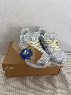 Asics silverfärgade sneakers - Snygga Asics sneakers i silver och vitt med mesh- och syntetmaterial. Skorna har krämvita snören, klassisk Asics-logga på sidan och Ortholite-innersula för extra komfort. Perfekta för dig som gillar sportig och trendig stil.