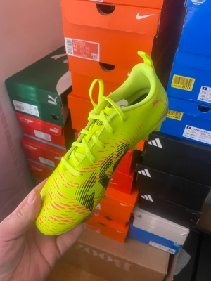 Neongula Puma fotbollssko - Snygg neongul fotbollssko från Puma med svarta och rosa detaljer. Skon har klassisk snörning och är tillverkad i ett lätt syntetmaterial för bra bollkontroll och snabbhet på planen. Perfekt för dig som vill sticka ut på fotbollsplanen.