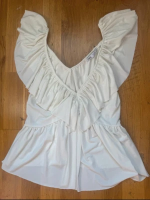 Vit volangtop från Zara - Säljer en söt vit topp från Zara med djup V-ringning och dubbla volangdetaljer framtill. Toppen har korta ärmar och ett lätt, luftigt tyg som ger en flowy känsla. Perfekt för att skapa en romantisk och trendig look.