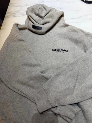 Grå Essentials Fear of God hoodie - Säljer en stilren grå hoodie från Essentials Fear of God. Hoodien har en klassisk huva och tryckt logga på bröstet samt större logga på ryggen. Tillverkad i mjukt bomullsmaterial som känns skönt mot huden. Perfekt för dig som gillar streetwear och enkel design.