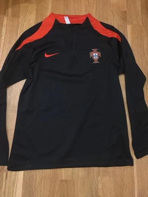 Portugal Nike långärmad träningströja - Snygg svart långärmad träningströja från Nike med Portugals landslagsmärke på bröstet och röda detaljer på axlar och krage. Tröjan har halv dragkedja framtill och är tillverkad i ett lätt, funktionellt material som passar perfekt till fotbollsträning. Passar om du är 160 - 165 cm