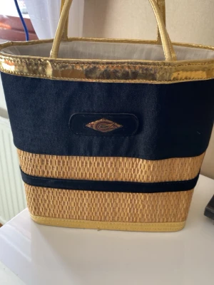 Svart och guld axelväska  - Snygg axelväska med svart tyg upptill och flätade strådetaljer nedtill. Väskan har guldfärgade handtag och kant, samt en dekorativ logga framtill. Perfekt för dig som vill sticka ut med en unik accessoar.