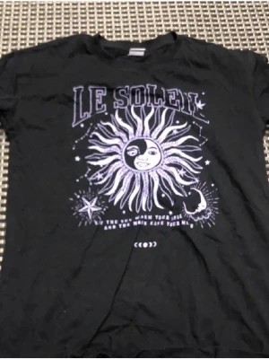 Svart t-shirt med soltryck från Lindex - Cool svart t-shirt från Lindex med ett stort tryck av en sol och texten 'Le Soleil' i lila och vitt. T-shirten har korta ärmar och är gjord i mjuk bomull. Perfekt för dig som gillar unika prints och vill sticka ut lite extra.