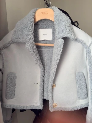 Ljusblå teddyjacka från Bershka - Supermjuk ljusblå teddyjacka från Bershka med stora fickor framtill och bred krage. Jackan har en croppad passform, synliga sömmar och stängs med knappar. Perfekt för dig som vill ha en cozy och trendig look.