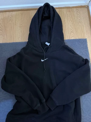 Svart Nike hoodie med huva - Säljer en svart hoodie från Nike med klassisk huva och dragsnören. Tröjan har en enkel design med Nike-loggan broderad i vitt på bröstet. Tillverkad i mjukt material som passar perfekt för en avslappnad stil.