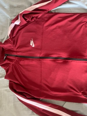 Röd Nike street jacket  - Snygg röd träningsjacka från Nike med vita ränder längs ärmarna och vit Nike-logga på bröstet. Jackan har hel dragkedja framtill, ståkrage och fickor med dragkedja. Perfekt för träning eller chill. Materialet är troligtvis polyester för en sportig känsla.