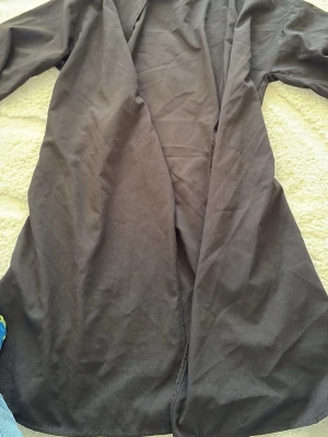 Svart lång kappa från Tavin - Stilren svart lång kappa från Tavin i ett tunt, mjukt tyg. Öppen framtill utan knappar eller dragkedja, vilket ger en avslappnad och modern look. Perfekt att slänga över en outfit för extra lager och stil.