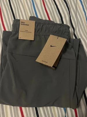Grå träningsbyxor från Nike Unlimited - Snygga grå träninggbyxor från Nike Unlimited med elastisk midja och dragsko. Byxorna har flera praktiska fickor på sidorna och är tillverkade i ett lätt och smidigt material som passar perfekt för en aktiv vardag. Stilsäker och funktionell design.