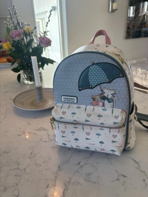 Söt ryggsäck med kattmotiv från Sammao - Liten och gullig ryggsäck i vitt och ljusblått från Sammao. Framsidan har ett tryck med två katter under ett paraply och små paraplymönster över hela väskan. Ryggsäcken har rosa handtag, justerbara axelband och en praktisk framficka med dragkedja.