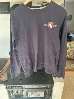 Svart långärmad topp från Gant - Säljer en svart långärmad topp från Gant med rund halsringning. På bröstet finns ett broderat Gant-emblem med röda och vita ränder samt texten '1949' i gult. Tillverkad i mjuk bomull, perfekt för en avslappnad och stilren look.