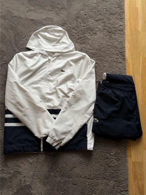 Lacoste Tracksuit - Snyggt träningsset från Lacoste med vit jacka och marinblå byxor. Jackan har huva, dragkedja och vita samt marinblå detaljer. Byxorna är marinblå med vita ränder på sidorna och har resår i midjan och vid bensluten. Båda plaggen har Lacostes klassiska krokodillogga. Passar M också