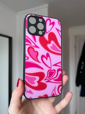 Y2K mobilskal med rosa hjärtan (iPhone 13 Pro Max) - Ett y2k-vibe mobilskal till iPhone 13 Pro Max med rosa bakgrund med röda och rosa hjärtan. Svarta kanter. Sparsamt använt.