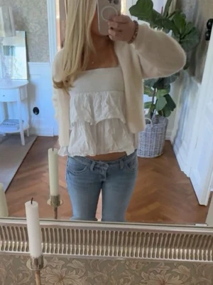 Vit volangig ärmlös topp - Säljer en söt vit topp med volanger och ärmlös design. Toppen har en croppad passform och raka axelband, perfekt för sommarens alla tillfällen. Tillverkad i ett lätt och luftigt bomullstyg som känns skönt mot huden.