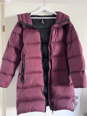 Lång vinröd dunjacka Pernilla Wahlgren - Snygg lång vinröd dunjacka från Pernilla Wahlgren med stor huva och svarta dragkedjor. Jackan har två stora fickor framtill. Perfekt för kalla vinterdagar och riktigt varm, bekväm och fin.💕Den är använd 1 gång därav väldigt bra skick.