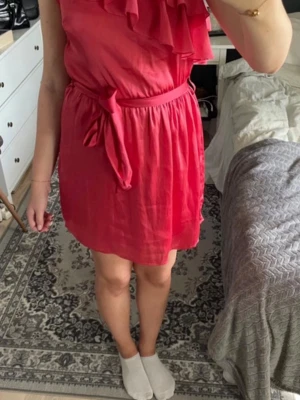 Rosa axelbandslös klänning med volang - Snygg rosa klänning med volangdetalj upptill och knytband i midjan. Axelbandslös modell med lätt utställd kjol och mjukt, tunt tyg som ger ett fint fall. Perfekt för dig som vill ha en feminin och trendig look.