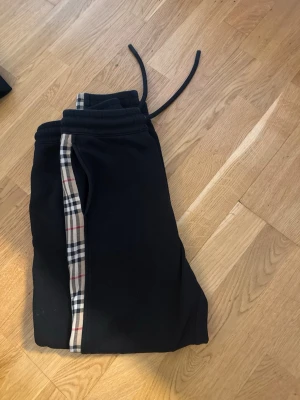 Svarta mjukisbyxor och hoodie med rutiga detaljer - Säljer ett par svarta mjukisbyxor och hoodie från Burberry med den klassiska beige, röda och svarta rutiga detaljen längs sidorna. Byxorna har dragsko i midjan och är gjorda i mjukt bomullsmaterial. Perfekta för en chill och stilren look.
