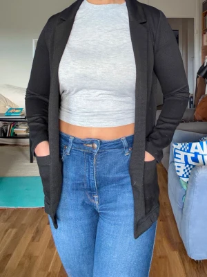 Svart kavaj med fickor - En stilren svart kavaj med klassisk krage och två framfickor. Kavajen har en rak passform och är tillverkad i ett mjukt, lätt material som passar perfekt till jeans eller mer dressade outfits. Enkel design utan knappar framtill. Passar från XS till M